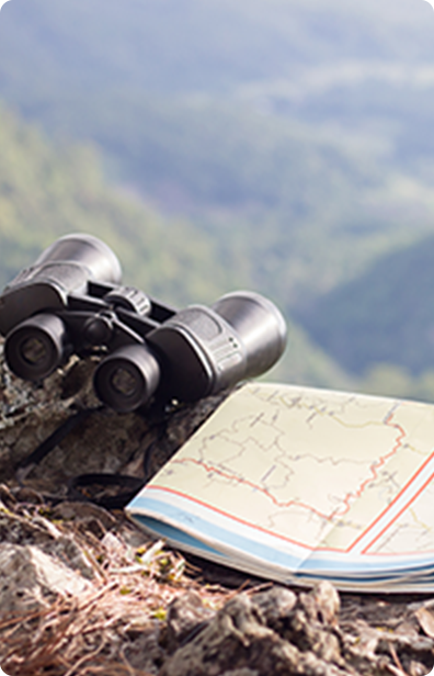 Binoculars and a folded map rest on a rocky surface outdoors, with a blurred landscape in the background.