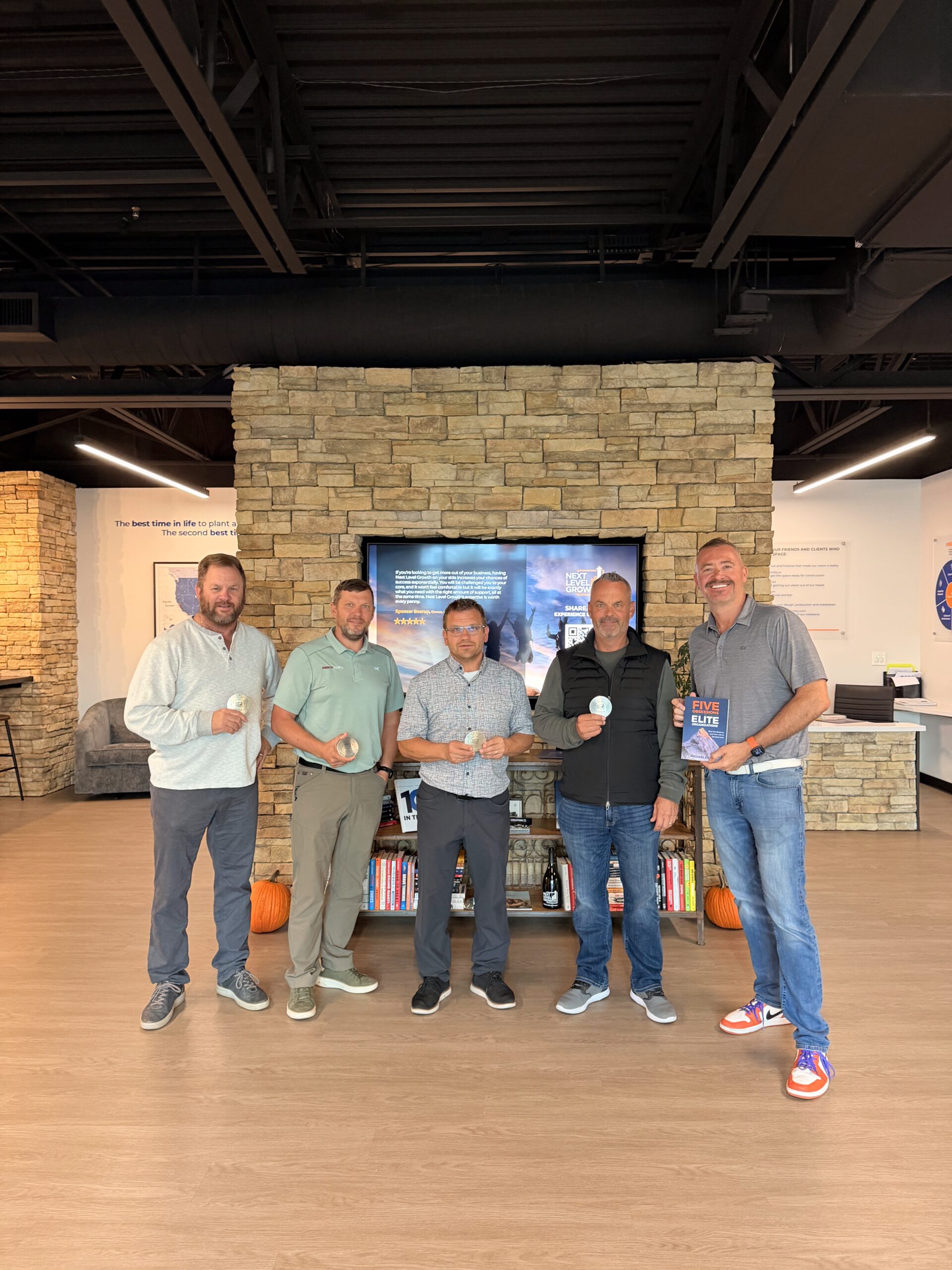 Five business coaches stand indoors in front of a stone fireplace and TV, each holding a round object, with books and pumpkins visible on the shelf below.