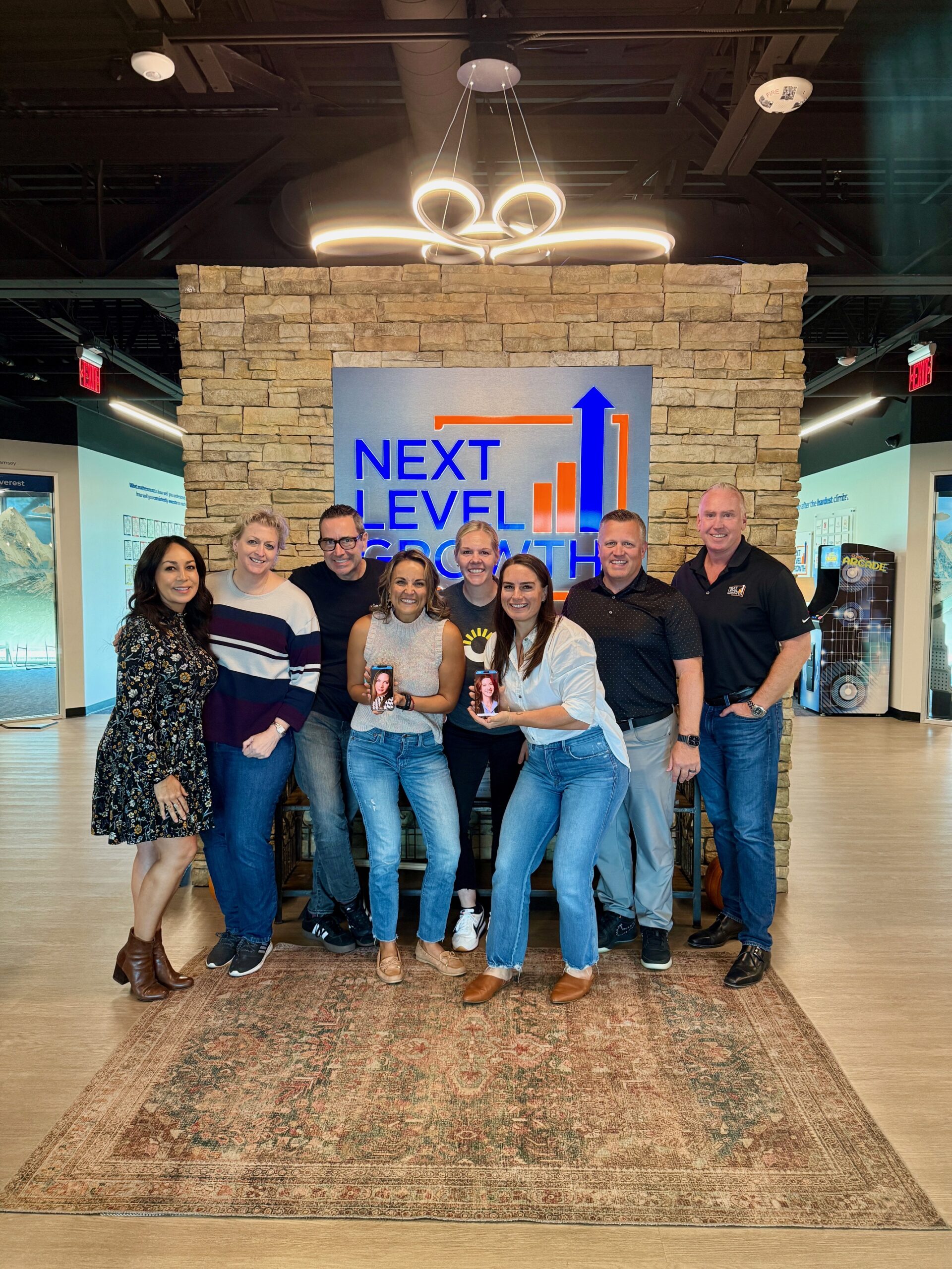 Eight people, including business coaches, stand smiling in front of a stone wall with a "Next Level Growth" sign in an office setting; some hold drinks and everyone is casually dressed.