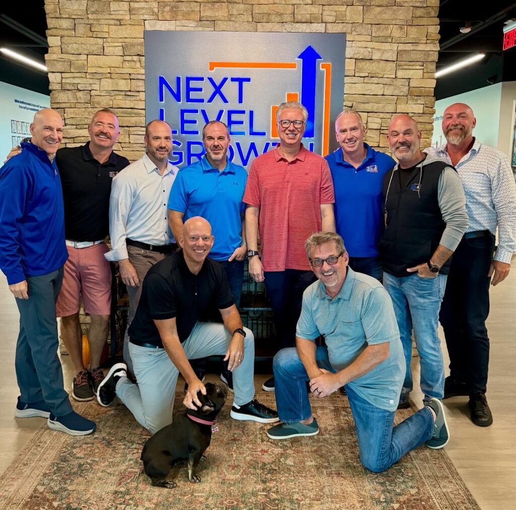 A group of ten men, acting as Business Coaches and Guides, pose and smile for a photo indoors in front of a "Next Level Growth" sign, with one man kneeling beside a small black dog.