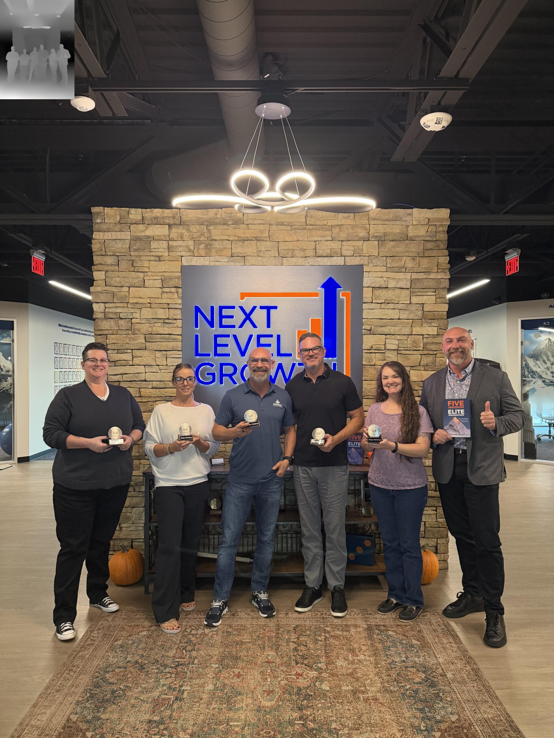 Six business coaches stand in front of a stone wall with a "Next Level Growth" sign, each holding an award, inside a modern office space.