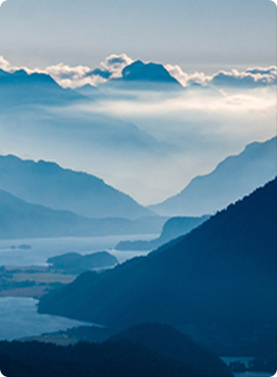 Blue mountains and hills layered in mist, with a river winding through the landscape under a cloudy sky.