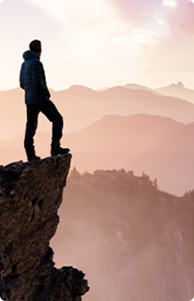 A person stands on the edge of a rocky cliff overlooking hazy, layered mountains at sunrise or sunset.