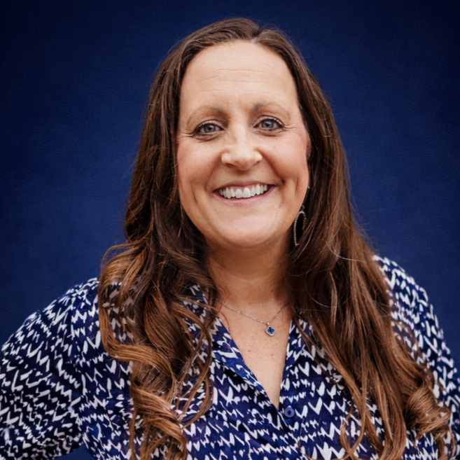 A woman with long brown hair wearing a blue and white patterned blouse stands in front of a solid dark blue background, smiling at the camera.