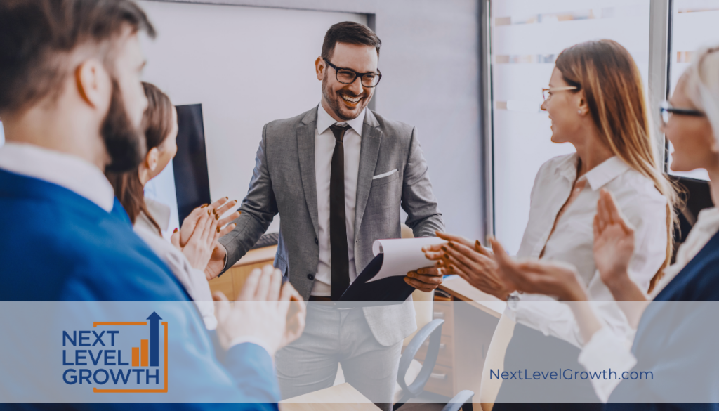 A man in a suit smiles while holding papers, standing with colleagues who are applauding in a modern office&mdash;celebrating success and fostering a high-performance culture. "Next Level Growth" logo and website appear on the image.