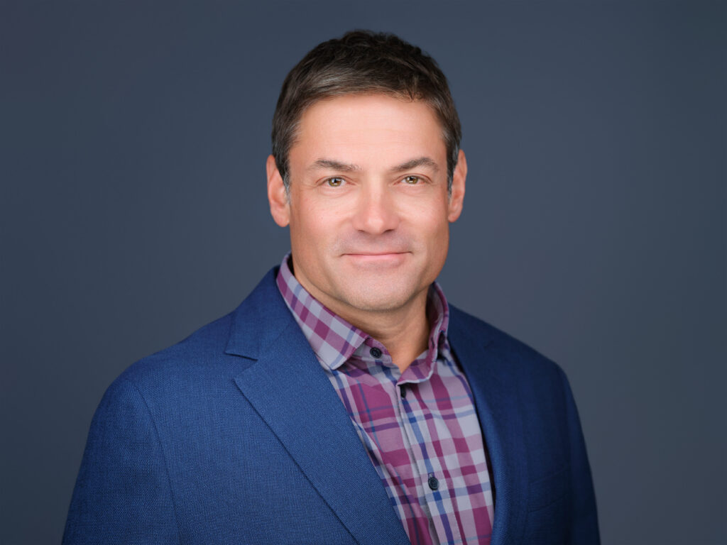 Man with short brown hair wearing a blue blazer over a plaid shirt, smiling slightly, posed against a plain dark background—this is David Ibanez.